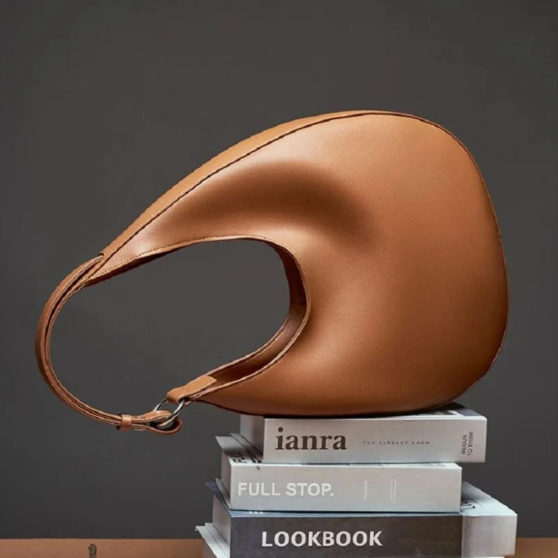 Display of an elegant brown leather bag, with a moon design and adjustable strap on stacked books.