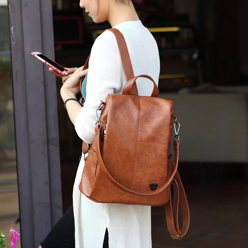 A stylish brown anti-theft leather backpack carried by a woman in a casual outfit.