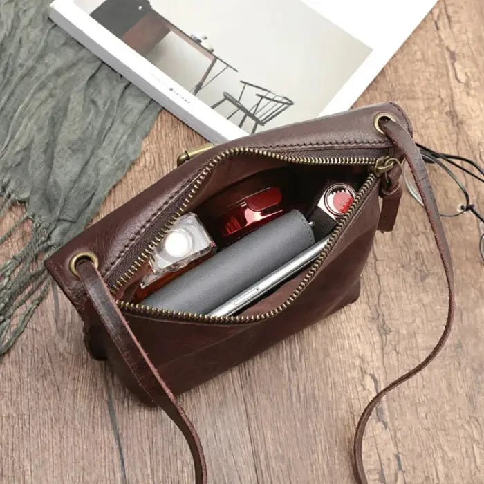 an open brown leather crossbody bag, showing perfume, a notebook and a cell phone inside.