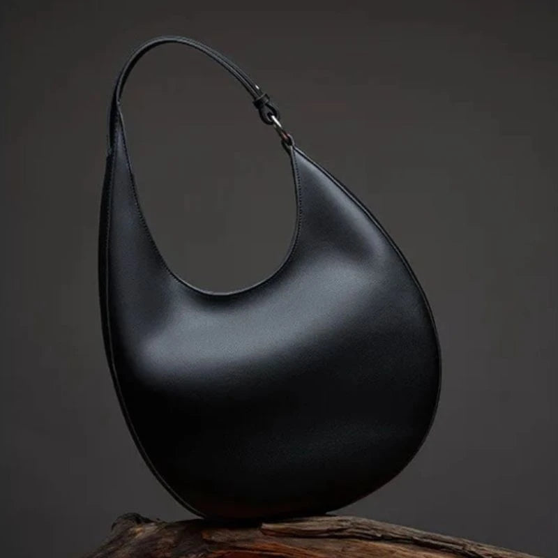 Display of an elegant black leather bag, with a moon design and adjustable strap, on a wood branch.