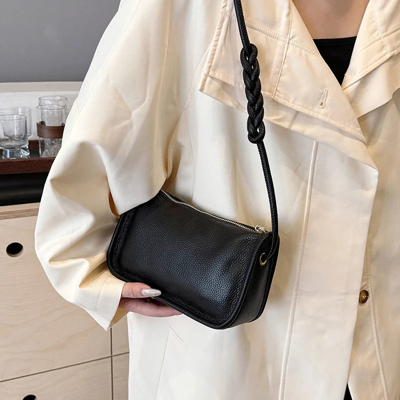 A stylish black leather crossbody bag with a braided strap, held on shoulder by a person in a cream-colored jacket.