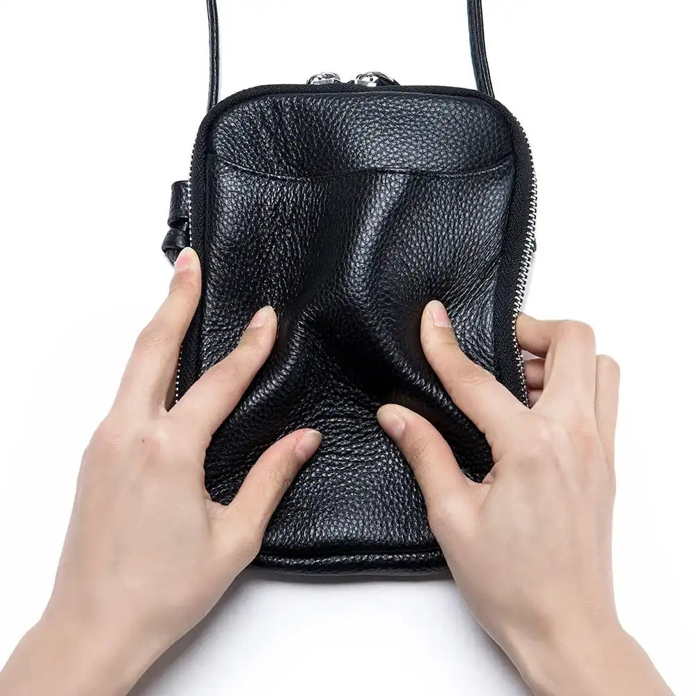 a black leather crossbody bag being squeezed with hands showing the flexibility of the material.