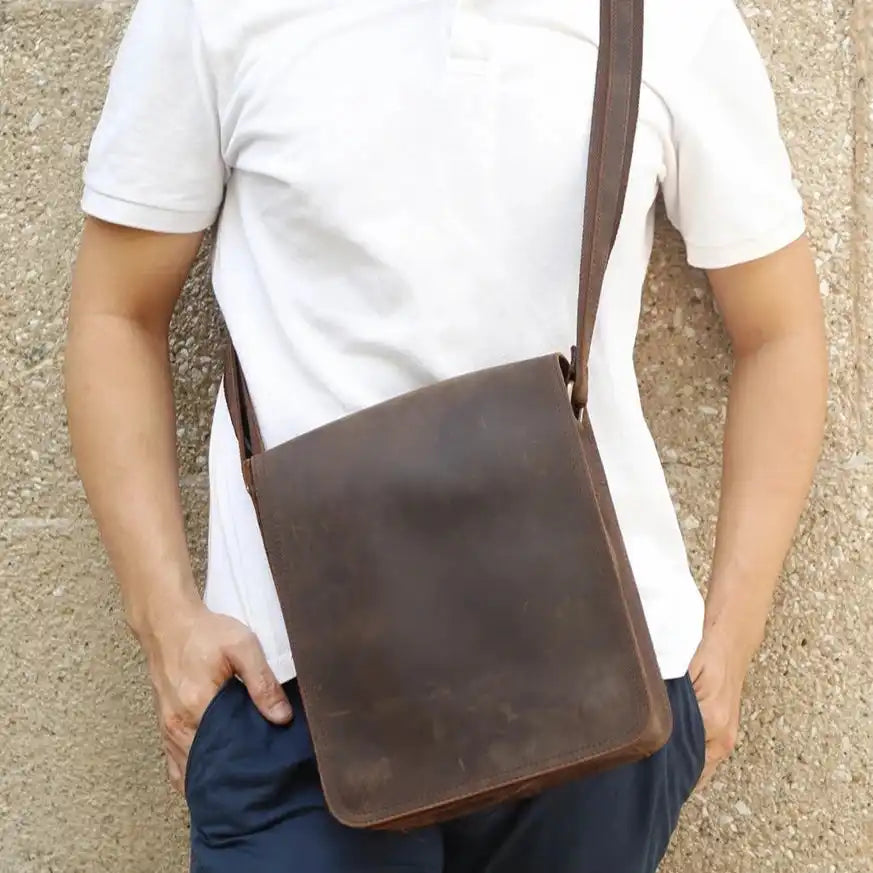 A man wearing a minimalistic brown leather crossbody bag over his shoulder, posing in front of a stone wall.