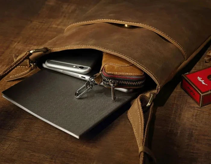 open leather crossbody bag revealing a smartphone, wallet, notepad on wooden table.
