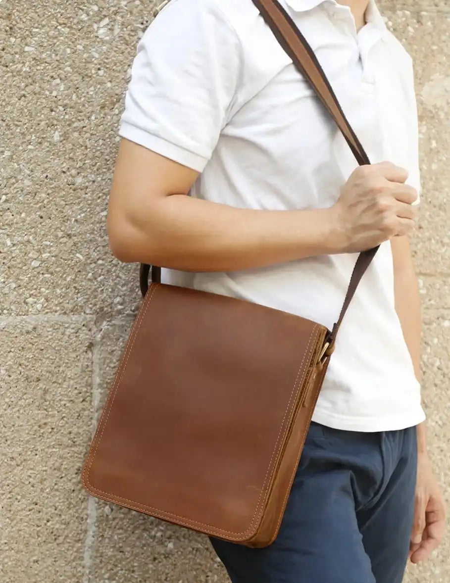 A man holding a brown leather crossbody bag with a white t-shirt. The bag displays a sleek and minimalistic design.