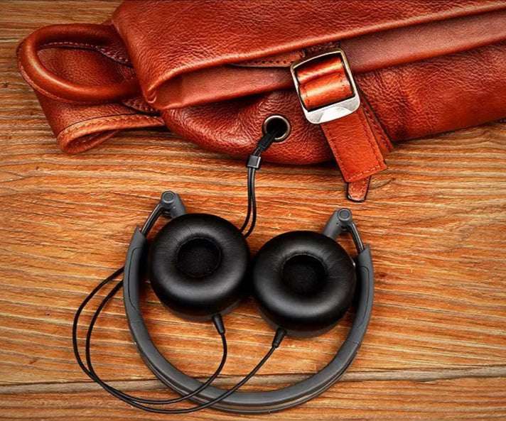 A pair of black headphones lying beside a brown leather bag on a wooden background.