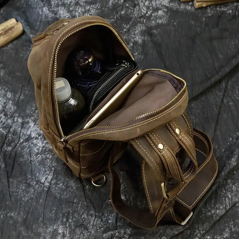 Open brown leather sling bag with internal compartments, showing water bottle, umbrella and a mobile device. 