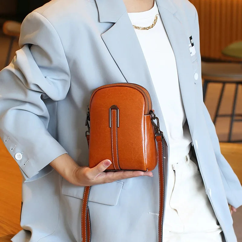 Front view of a stylish caramel color leather crossbody bag held by a person in a light blue blazer.