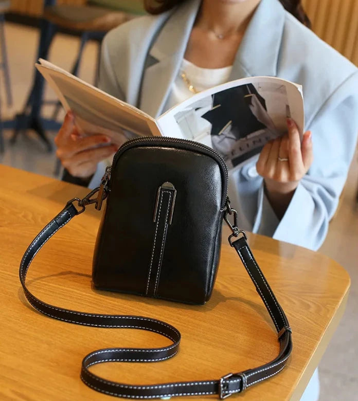 A stylish black leather crossbody bag on a table with a woman reading a magazine.