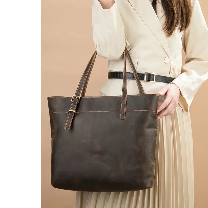 A stylish brown leather tote bag held by a woman in a cream-colored outfit.