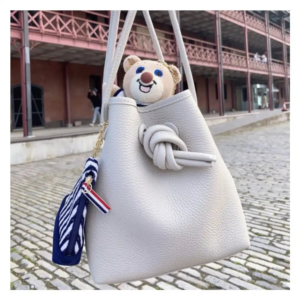 A plush bear toy sitting inside a chic leather white handbag with knot design on a cobblestone street.