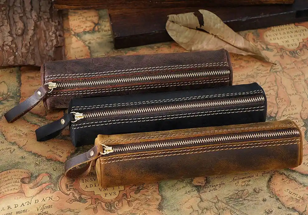 display of three leather pen cases in brown, black and coffee brown. The cases have zippers, handles and stitching details. 