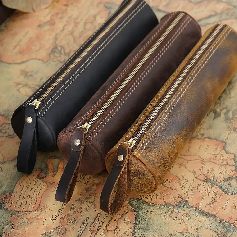 display of three leather pen cases in brown, black and coffee brown. The cases have zippers and stitching details. 