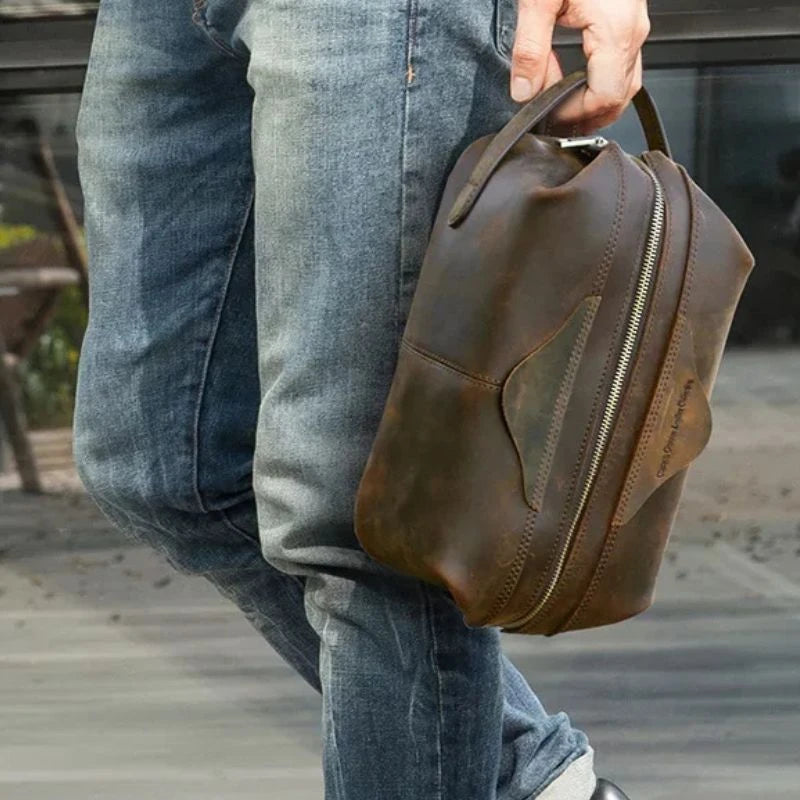 A man in blue jeans holding a brown leather toiletry bag. 