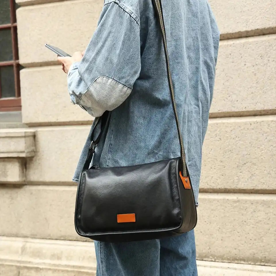 A person wearing a denim shirt holding a smartphone, while carrying a black leather bag on the shoulder.