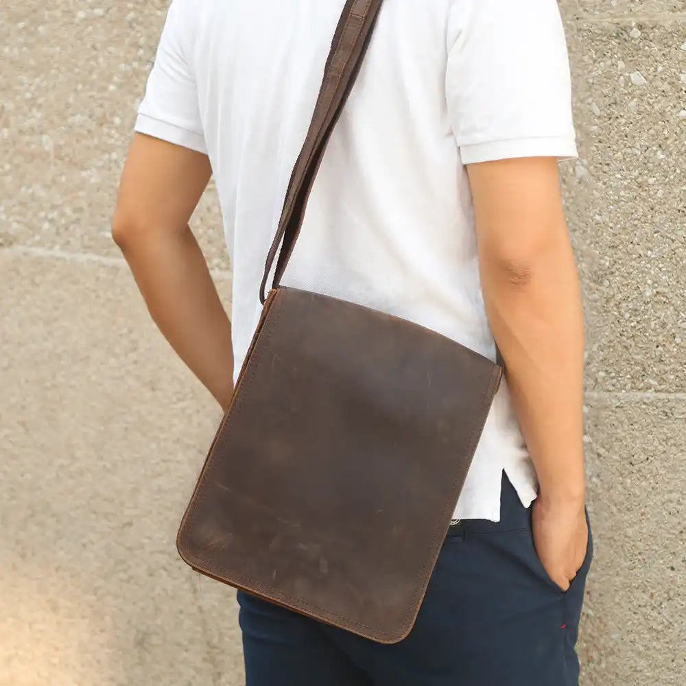 A man with a brown leather crossbody bag over his shoulder, wearing a white t-shirt posing in front of a stone wall.