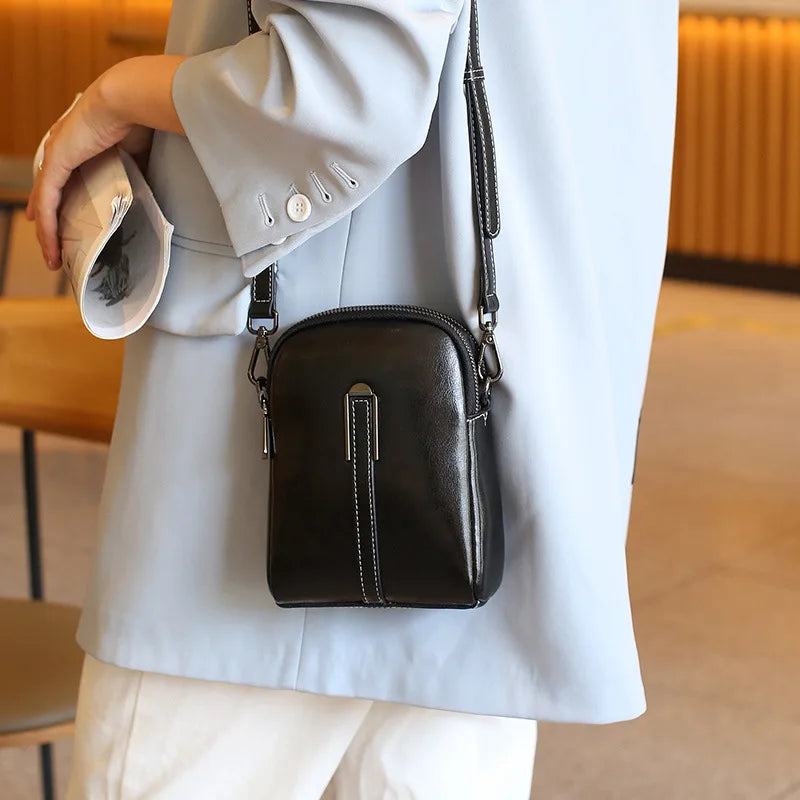 A fashionable black leather crossbody bag worn across the body by a person wearing a light blue coat showcasing contemporary style.