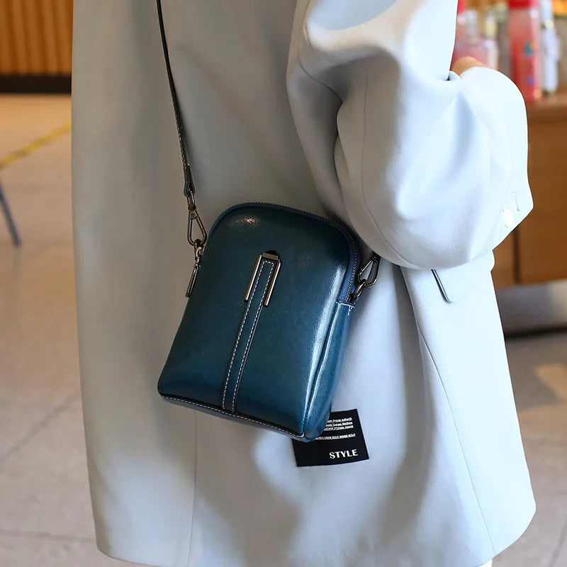 A modern blue leather crossbody bag with a sleek design, hanging from a shoulder.