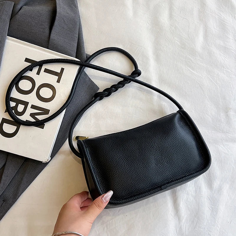 A chic black crossbody bag with braided strap held in a hand, laid on a textured surface.