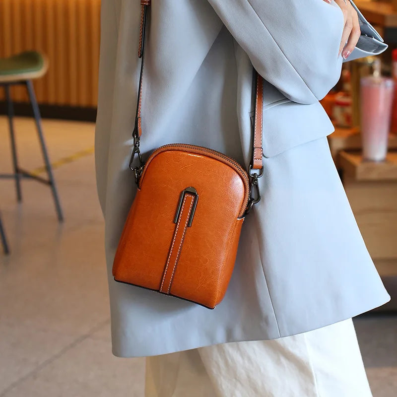 A stylish caramel color leather crossbody bag worn by a person in a light blue outfit.