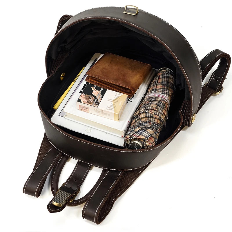 Open dark brown leather backpack, displaying books, a notebook, an umbrella and a wallet showcasing large capacity.